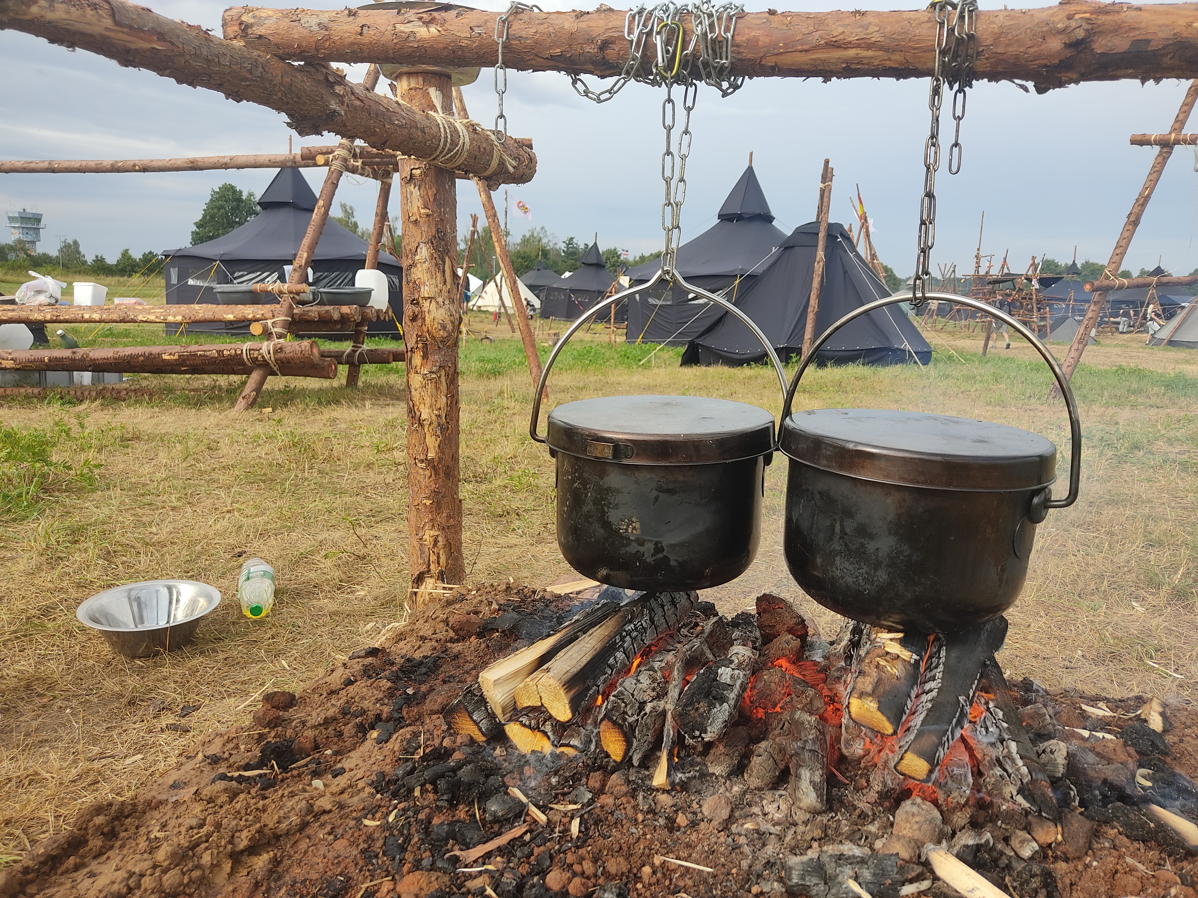 Zwei gro&szlig;e Kocht&ouml;pfe h&auml;ngen &uuml;ber einem Lagerfeuer, im Hintergrund das Camp